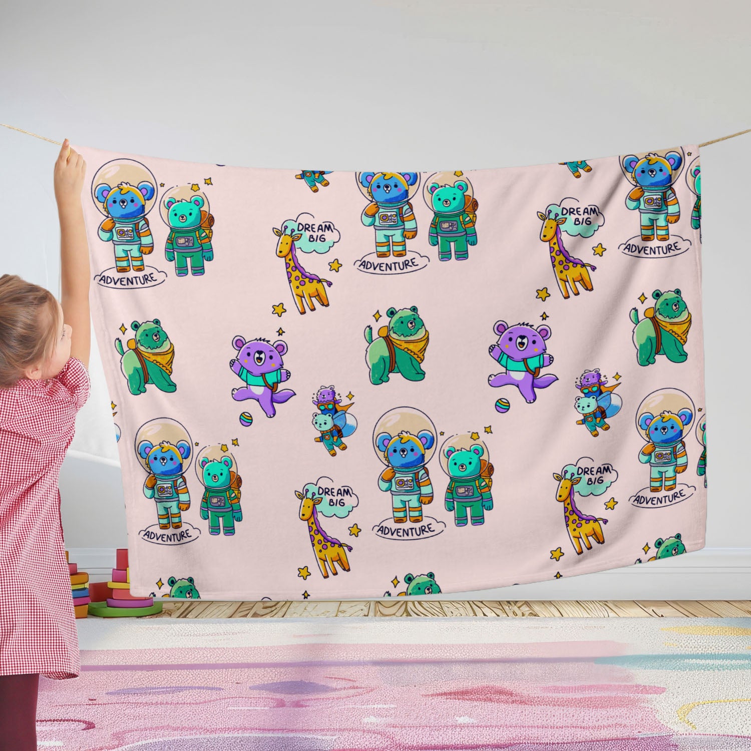Child holding up a pastel-pink throw blanket printed with cute cartoon astronaut bears, giraffes, and “Dream Big Adventure” text in a bright playroom setting.