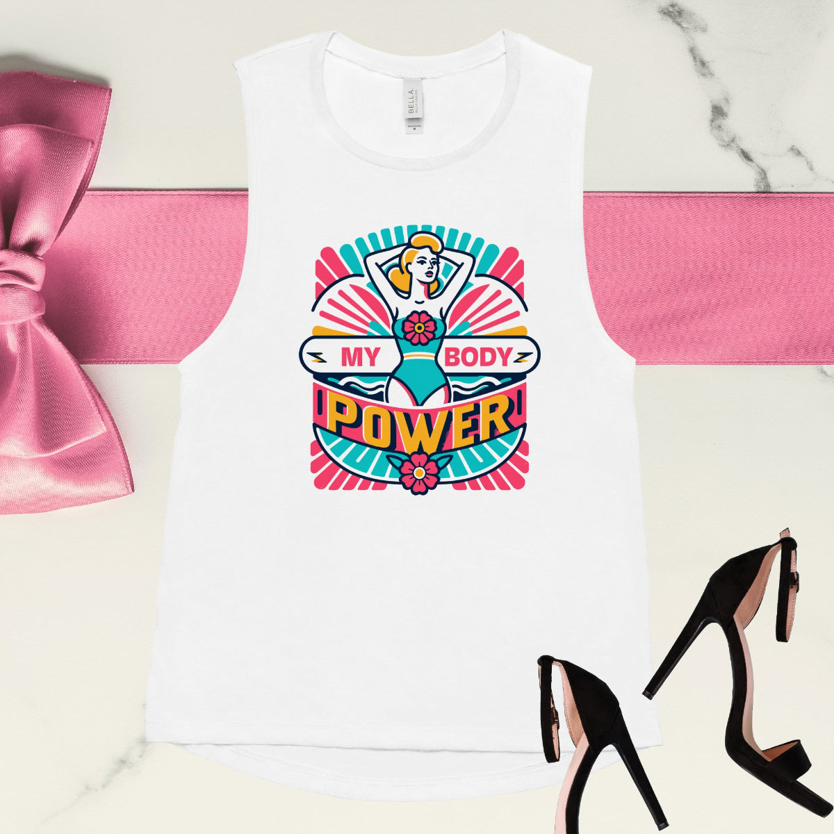 A white muscle tank top with a vibrant graphic design of a woman and the text 'MY BODY POWER' on the front, presented on a marble surface with a pair of black high-heeled shoes and a pink bow to the side.