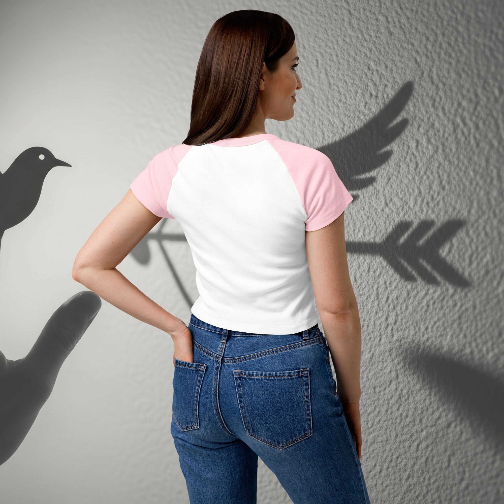 Woman wearing a white and pink shirt with blue jeans, standing against a gray wall with shadow effects.