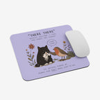 A lavender rectangular mouse pad with rounded corners, featuring a printed illustration of a black cat and a robin bird with the text “THERE THERE” and a quote. A white computer mouse rests on the right side of the pad against a light gray background.