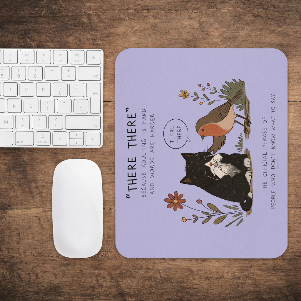 A rectangular desk mat with rounded corners sits on a wooden tabletop next to a white keyboard and mouse. The mat has a lavender background with a black-and-white cat, a robin bird, flowers, and the phrase “THERE THERE” printed on it.