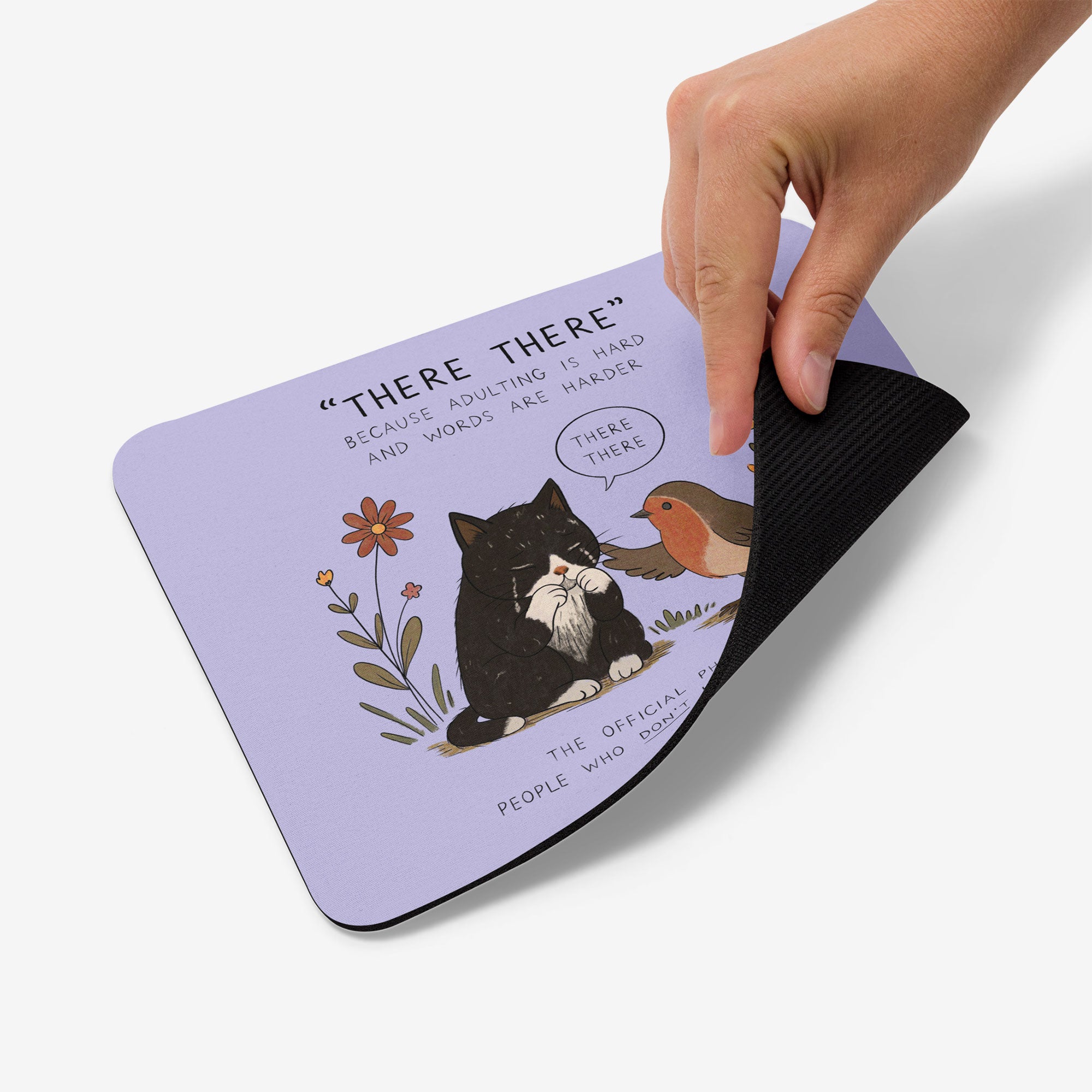 A hand is lifting the corner of a lavender mousepad to show its black textured underside. The top surface features a cute illustration of a black cat and a robin bird with the text 'THERE THERE BECAUSE ADULTING IS HARD AND WORDS ARE HARDER THERE THERE'.