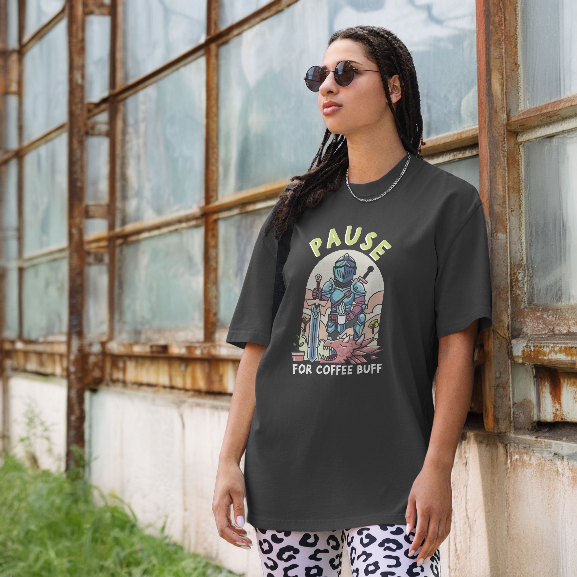 Person wearing a graphic t-shirt with a knight design and text pause for coffee buff, standing in front of an industrial old building.