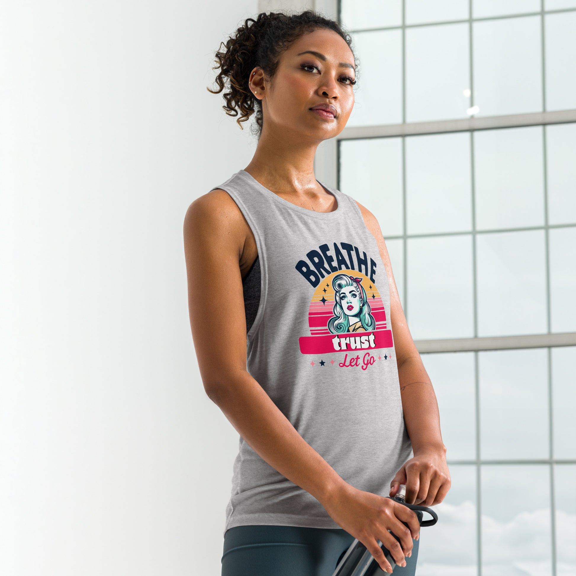 Woman wearing Ladies’ Muscle Tank with "Breathe, Trust, Let Go" graphic, holding resistance bands in a bright, airy gym setting.